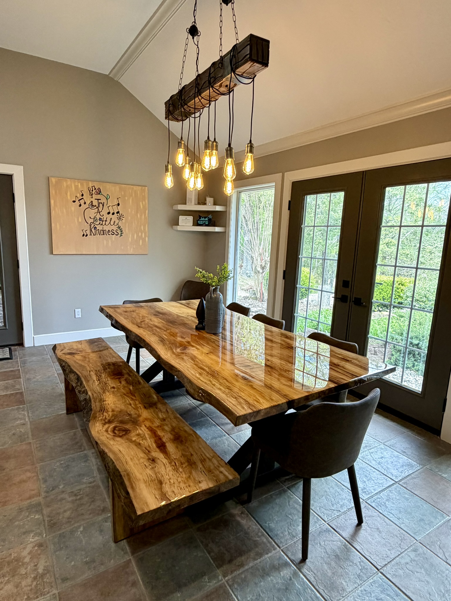 Sycamore dining table closeup with matching bench Edison bulb chandelier and Try a Little Kindness art